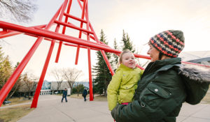 A woman holds a child in her arms while standing near a striking red and very large artwork, forming an archway across the walking path.